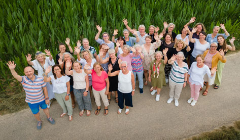 Das Bild zeigt eine große Gruppe von Frauen und einen Mann, die vor einem hohen Grasfeld stehen und alle mit erhobenen Händen winken. Sie scheinen eine Gruppe von Menschen zu sein, die sich gut kennen und gemeinsam etwas feiern oder ein gemeinsames Ereignis haben. Die Stimmung ist fröhlich und positiv. Die Kleidung der Personen ist leger und die Farben sind vielfältig und bunt. Das Bild vermittelt einen Eindruck von Gemeinschaft und Zusammengehörigkeit.