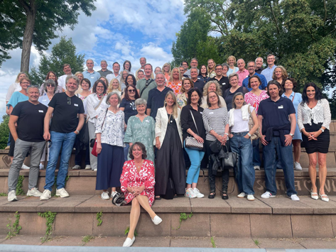 Eine große Gruppe erwachsener Menschen steht und sitzt auf Steinstufen vor Bäumen und einem bewölkten Himmel.
