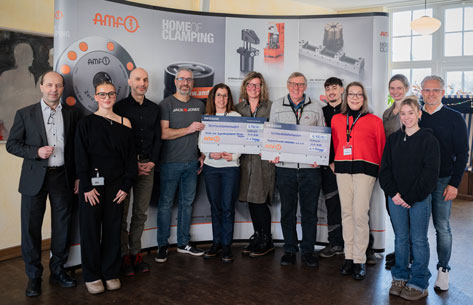 Eine Gruppe von etwa einem Dutzend Menschen steht vor einem Werbebanner und hält symbolische Schecks.  Im Detail zeigt das Bild eine Gruppe von elf Personen, die sich vor einem Firmenbanner in einem Innenraum versammelt haben. Das Banner ist mit dem Logo 