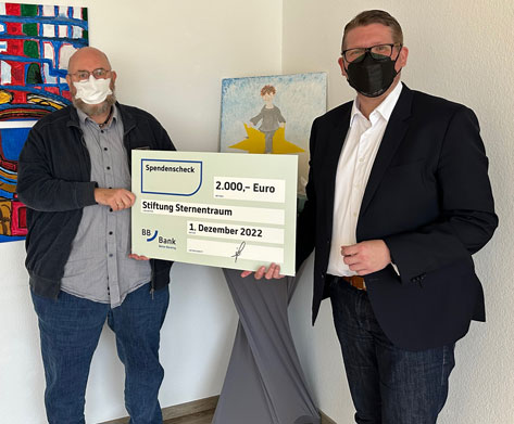 Vorstand Jürgen Müller und Herr Bringe von der BBBank stehen mit einem großen Spendenscheck vor einem bunten Bild.