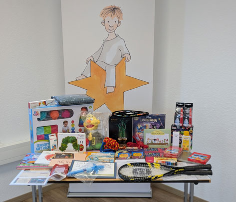Auf einem Tisch sind verschiedene Geschenke und Spielzeuge arrangiert. Im Hintergrund ist eine Zeichnung eines Kindes zu sehen, das auf einem Stern sitzt. Die Geschenke umfassen unter anderem ein sensorisches Geschenkset, Bücher, Spiele wie 