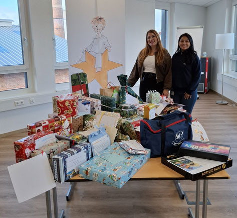 Das Bild zeigt zwei Frauen, die vor einem Tisch voller Geschenke stehen. Im Hintergrund ist ein Banner mit einer Zeichnung eines Kindes zu sehen, das auf einem Stern sitzt. Die Geschenke sind in verschiedenfarbiges Geschenkpapier eingepackt und scheinen für einen wohltätigen Zweck oder eine Weihnachtsaktion zu sein. Auf dem Tisch stehen außerdem eine blaue Tasche und ein Set Acrylfarben.