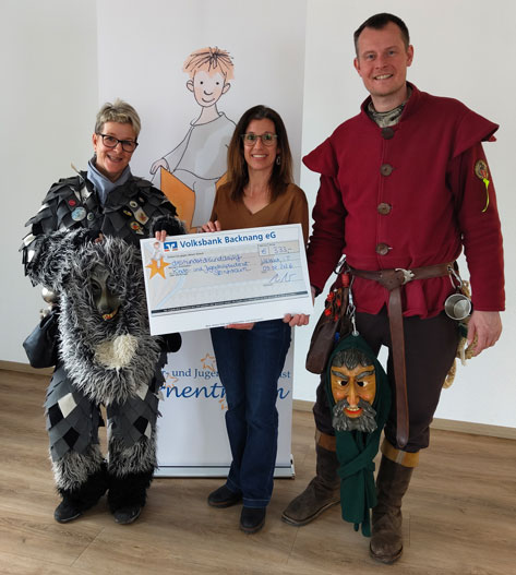 Drei Personen stehen vor einem Banner und präsentieren einen übergroßen Scheck. Im Detail zeigt das Bild drei Personen, die vor einem Banner posieren und einen übergroßen Scheck der Volksbank Backnang eG präsentieren. Die Szene wirkt wie eine Spendenübergabe oder eine ähnliche Ehrung. Die Person links trägt ein auffälliges, dunkles Faschingskostüm, das an ein Zotteltier erinnert. Die Frau in der Mitte trägt eine braune Bluse und Jeans, wirkt formell und hält den Scheck mit. Der Mann rechts ist ebenfalls in einem traditionellen Faschingskostüm gekleidet, einschließlich einer roten Jacke und einer Maske, die an seinem Gürtel hängt. Der Hintergrund wird von einem Banner mit einer Illustration eines Kindes und einem Schriftzug dominiert, der auf ein Kinder- und Jugendhospizdienst Sternentraum hinweist. Der Boden ist hell und reflektierend, was auf eine polierte Oberfläche hindeutet. Die Atmosphäre ist positiv und festlich, was durch die ungewöhnlichen Kostüme und den übergroßen Scheck unterstrichen wird.