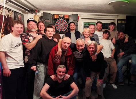 Dieses Foto zeigt eine Gruppe von Personen unterschiedlichen Alters, die sich in einem gut beleuchteten Raum versammelt haben, wobei im Hintergrund eine Dartscheibe zu sehen ist.  Die Szene ist im Querformat aufgenommen und die Gruppe steht eng zusammen, was ein Gefühl der Gemeinschaft und Vertrautheit vermittelt. Die Komposition ist leicht asymmetrisch, mit einer dichteren Ansammlung von Personen auf der linken und mittleren Seite des Bildes. Im Vordergrund kniet eine Person, während die anderen hauptsächlich stehen und sich teils umarmen oder die Arme um Schultern legen. Die Körpersprache ist insgesamt entspannt und positiv, was auf eine lockere Zusammenkunft hindeutet. Der Blickwinkel ist etwa auf Augenhöhe der meisten stehenden Personen, was eine direkte und persönliche Perspektive schafft.  Die Personen auf dem Bild variieren in Alter und Geschlecht. Viele der Männer tragen Freizeitkleidung wie T-Shirts, Hoodies und Jeans. Ein Mann im Vordergrund, der kniet, hat einen kurz geschorenen Kopf und einen freundlichen Gesichtsausdruck, während er seine Hände vor sich gefaltet hat. Der Mann links daneben trägt ein schwarzes T-Shirt und scheint eine blonde Frau neben sich zu umarmen. Weitere Personen im mittleren Hintergrund zeigen unterschiedliche Gesichtsausdrücke, von Lächeln bis hin zu neutralen Blicken. Die Altersspanne reicht von jüngeren Erwachsenen bis hin zu älteren Personen. Kleidung und Frisuren sind vielfältig, aber sie alle scheinen Teil derselben informellen Gruppe zu sein.  Das Foto ist eine digitale Fotografie. Der Stil ist realistisch und ungestellt, typisch für eine Momentaufnahme einer privaten Feier oder eines Treffens. Die technische Ausführung ist solide, mit guter Belichtung und Schärfe, die es ermöglicht, die Gesichter und Details der Personen gut zu erkennen. Es gibt keine offensichtlichen künstlerischen Effekte oder Filter, was dem Bild eine authentische und ungekünstelte Atmosphäre verleiht. Der Fokus liegt klar auf den Personen und ihrer Interaktion.  Der Raum, in dem sich die Gruppe befindet, scheint ein privater Aufenthaltsraum oder eine Bar zu sein. An den Wänden hängen Poster, darunter eines, das wie ein Rock-Album-Cover aussieht. Eine Dartscheibe mit Pfeilen ist prominent im Hintergrund platziert, was auf Freizeitaktivitäten hindeutet. Die Beleuchtung ist hell und gleichmäßig, wahrscheinlich von Deckenleuchten, was die Szene gut ausleuchtet und keine dramatischen Schatten erzeugt. Die Umgebung wirkt gemütlich und einladend, was die positive Stimmung der Gruppe unterstreicht. Die Wände sind in einem hellen Farbton gehalten, und im Hintergrund sind auch Türen oder Fenster zu erkennen.