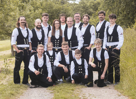 Das Bild zeigt eine Gruppe von etwa 15 Personen, die in traditioneller Zimmermannskleidung posieren. Die meisten tragen schwarze Westen mit Knöpfen, weiße Hemden und dunkle Hosen oder Lederhosen. Es sind sowohl Männer als auch Frauen und Kinder in der Gruppe. Sie stehen oder hocken auf einem Pfad in einer grünen, natürlichen Umgebung, wahrscheinlich im Freien. Der Hintergrund besteht aus Bäumen und Vegetation.