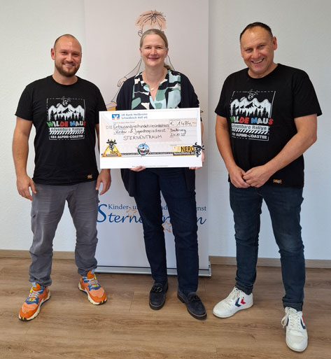 Das Bild zeigt drei Personen, die anscheinend eine Spende überreichen. In der Mitte hält eine Frau einen übergroßen Scheck über 1.484 Euro. Auf dem Scheck steht der Name der Spendenorganisation „Sternentraum“. Die Personen links und rechts von ihr sind männlich. Alle drei lächeln in die Kamera. Sie stehen vor einem Banner, auf dem das Logo der Organisation „Kinder- und Jugenddienst Sternentraum“ abgebildet ist. Die beiden Männer tragen T-Shirts mit der Aufschrift „Wilde Maus Adventure Rallye Alpine-Coaster“.