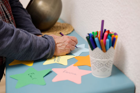 Das Bild zeigt die Hände einer Person, die mit einem lila Stift auf einen gelben Stern aus Papier schreibt. Auf dem Tisch liegen weitere bunte Sterne, einige bereits beschriftet mit Namen wie 