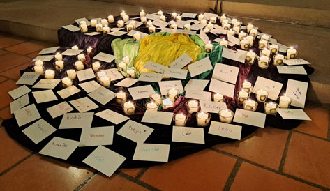 Das Bild zeigt ein Arrangement von Kerzen und Kärtchen mit Namen auf einem erhöhten Podest, das mit Stoffen in Lila, Grün und Gelb bedeckt ist. Die Kerzen, die in kleinen Glasbehältern stehen, sind über die gesamte Fläche verteilt und leuchten, während die Kärtchen mit verschiedenen Namen in unterschiedlichen Schriftarten und Farben beschriftet sind und um die Kerzen herum platziert wurden. Der Untergrund besteht aus einem Fliesenboden. Die Szene wirkt wie ein Gedenkort oder eine Zeremonie, in der einzelnen Personen gedacht wird.